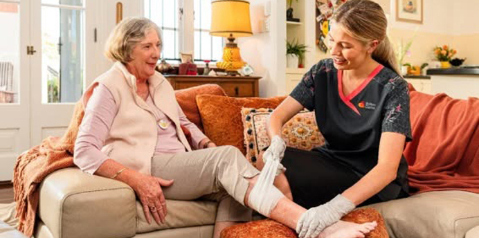 A healthcare worker applies a bandage to an older person's leg while both are seated in a living room